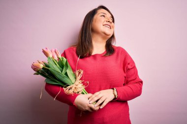 Güzel ve cüsseli bir kadın pembe arka planda romantik laleler tutuyor. Yüzünde gülümseme, doğal bir ifade ile yan yana bakıyor. Kendinden emin gülen.