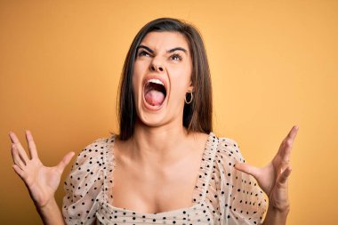 Sarı arka planda sıradan tişörtler giyen genç, güzel esmer kadın. Deli deli bağırıyor ve agresif bir ifadeyle bağırıyor ve kollarını kaldırıyor. Hayal kırıklığı konsepti.
