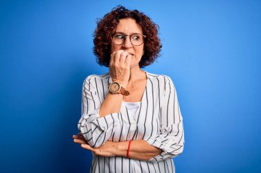 Orta yaşlı güzel kıvırcık saçlı kadın izole edilmiş arka planda sıradan çizgili tişört giyiyor. Elleri ağızdan tırnak yerken stresli ve gergin görünüyor. Endişe sorunu.