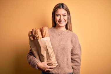 Genç, güzel bir kadın, elinde kağıt torbayla, izole edilmiş sarı arka planda, mutlu bir yüzü olan, kendine güvenen bir gülümsemeyle duran, dişlerini gösteren bir çanta tutuyor.