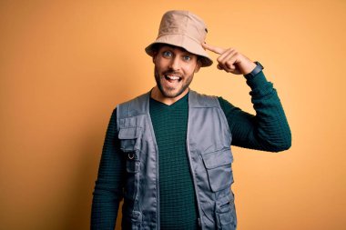 Tatilde sakallı yakışıklı turist sarı arka planda kaşif şapkası takıyor. Tek parmağıyla başını işaret ediyor. Harika bir fikir ya da düşünce, iyi bir hafıza.