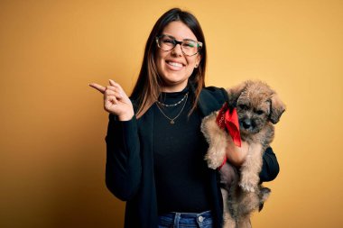 Genç, güzel esmer bir kadın, sevimli bir köpek yavrusunu izole edilmiş sarı arka planda tutarken mutlu bir şekilde elini ve parmağını kenarı işaret ediyor.