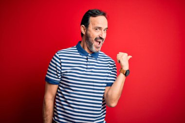 Orta yaşlı, beyaz çizgili polo giyen, izole edilmiş kırmızı arka planda dikilen mutlu yüzüyle gülümseyen ve baş parmağıyla yan tarafı gösteren bir adam..
