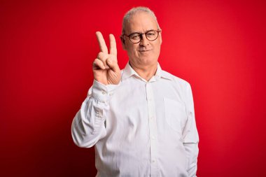 Orta yaşlı, beyaz gömlekli ve kırmızı arka planda gözlüklü yakışıklı adam kameraya bakıp zafer işareti yaparken gülümsüyor. İki numara..