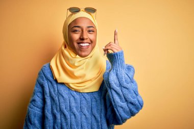 Sarı başörtüsü takmış, güneş gözlüklü genç Afrikalı Amerikalı bir bayan başarılı bir fikir işaret ediyor. Heyecanlı ve mutlu. Bir numara..