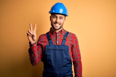 İnşaat üniforması ve sarı arka planda güvenlik kaskı takan genç müteahhit kameraya gülümseyip zafer işareti yapıyor. İki numara..