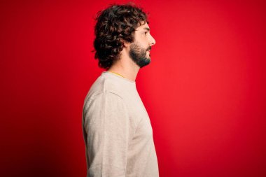 Sakallı, sıradan bir süveter giyen, kırmızı arka planda dikilen, rahat bir görünüm takınan, kendine güvenen bir gülümsemesi olan genç yakışıklı bir adam..