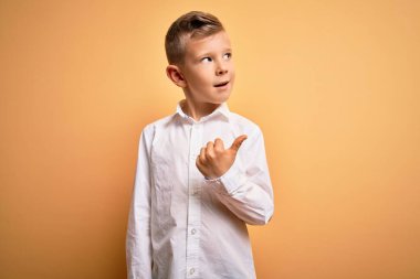 Küçük, beyaz, mavi gözlü, sarı arka planda zarif beyaz gömlek giyen mutlu yüzüyle gülümseyen ve baş parmağıyla yüzünü gösteren genç bir çocuk..