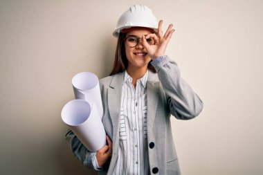 Young beautiful architect woman wearing safety helmet and glasses holding blueprints with happy face smiling doing ok sign with hand on eye looking through fingers