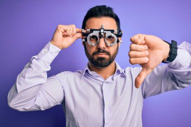 Mor arka planda optometri gözlüğü takan, sakallı, yakışıklı, yakışıklı, optik bir adam. Kızgın yüzlü, negatif işaretli, başparmakları aşağıda, reddedilmiş.