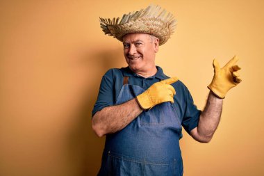 Sarı arka planda önlük ve şapka takan orta yaşlı çiftçi gülümseyip kameraya bakıyor ve iki eliyle ve parmaklarıyla yan tarafı işaret ediyor..