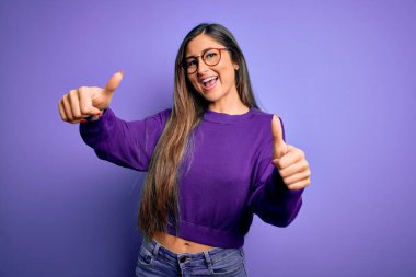 Mor izole edilmiş arka planda gözlük takan genç ve güzel bir kadın el hareketini onaylıyor, gülümsüyor ve başarı için mutlu. Kazanan jest.
