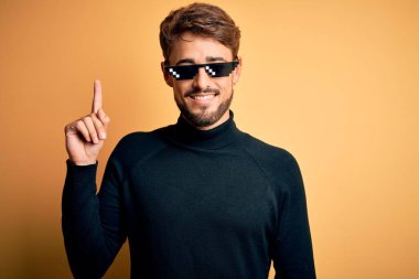 Haydut hayatı takan genç adam izole edilmiş sarı arka planda duran popo güneş gözlüğü başarılı bir fikirle işaret ediyor. Heyecanlı ve mutlu. Bir numara..