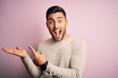 İzole edilmiş pembe arka plan üzerinde duran gündelik süveter giyen genç yakışıklı adam hayrete düşmüş ve kameraya gülümserken eliyle ve parmağıyla işaret ediyordu..
