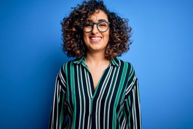 Güzel kıvırcık Arap kadın çizgili gömlek ve mavi arka planda gözlük takıyor ve yüzünde mutlu ve havalı bir gülümseme var. Şanslı kişi..