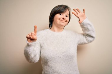 Genç Down sendromlu kadın izole edilmiş arka planda duruyor. Altıncı parmaklarını gösterip işaret ederken kendinden emin ve mutlu bir şekilde gülümsüyor..