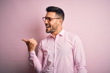 Zarif gömlekli ve gözlüklü genç adam pembe arka planda ayakta gülümsüyor, gülümsüyor ve başparmağıyla yana bakıyor..