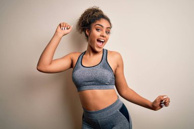 Genç Afrikalı Amerikalı spor kadın spor yapıyor, beyaz arka planda spor kıyafetleri giyiyor, mutlu ve neşeli dans ediyor, gülümsüyor, rahat hareket ediyor ve kendinden emin bir şekilde müzik dinliyor.