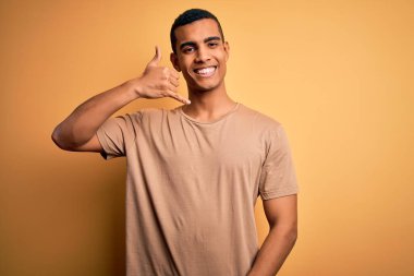 Genç, yakışıklı, Afro-Amerikalı bir adam sarı arka planda dikilmiş, rahat bir tişört giymiş telefonla el ve parmaklarıyla telefonla konuşuyor gibi gülümsüyor. İletişim kavramları.