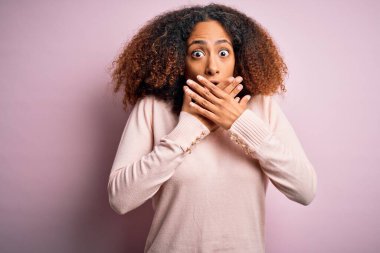 Afro saçlı, pembe arka planda günlük süveter giyen Afro-Amerikalı genç bir kadın yanlışlıkla ağızlarını kapatarak şok geçirdi. Gizli konsept.