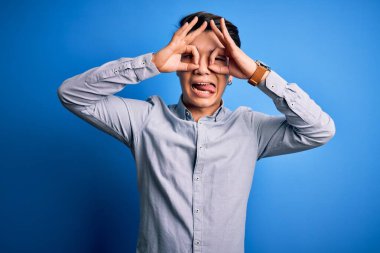 Genç yakışıklı Çinli bir adam, günlük gömleğini giymiş, mavi arka planda dikiliyor. Dürbün gibi güzel hareketler yapıyor. Gözleri parmaklarından bakıyor. Çılgın ifade.