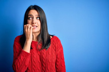Mavi gözlü genç esmer kadın izole edilmiş arka planda kırmızı bir süveter giyiyor. Elleri ağızdan tırnak yerken gergin ve stresli görünüyor. Endişe sorunu.