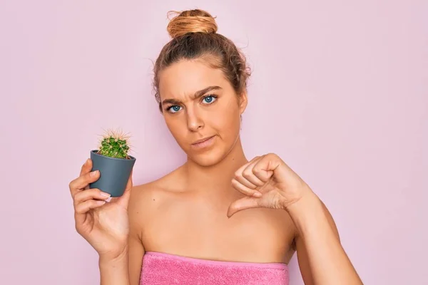 Beautiful blonde woman with blue eyes wearing towel shower after bath holding small cactus with angry face, negative sign showing dislike with thumbs down, rejection concept