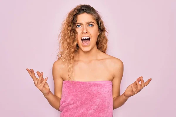Beautiful blonde woman with blue eyes wearing towel shower after bath over pink background crazy and mad shouting and yelling with aggressive expression and arms raised. Frustration concept.