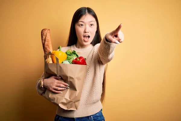 年轻的亚洲女人拿着一纸袋新鲜健康的食品放在黄色孤立的背景上指尖着前方的手指 张开嘴惊讶的表情 前面有什么东西 — 图库照片