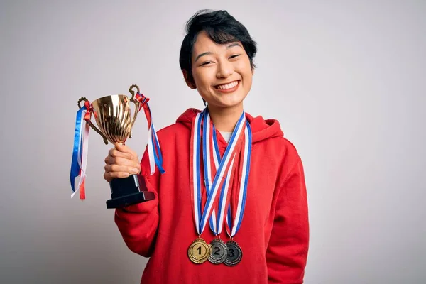 Young beautiful asian girl winner holding trophy wearing medals over white background with a happy face standing and smiling with a confident smile showing teeth