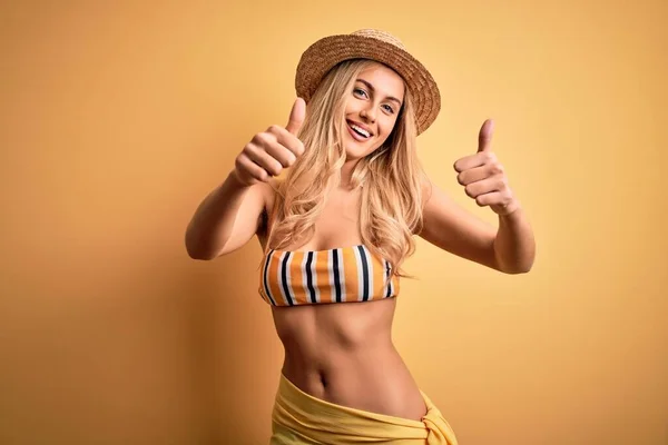 Young beautiful blonde woman on vacation wearing bikini and hat over yellow background approving doing positive gesture with hand, thumbs up smiling and happy for success. Winner gesture.