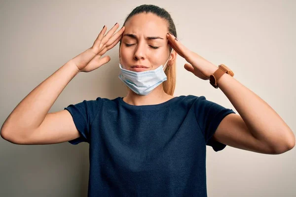 Beautiful blonde woman with blue eyes wearing medical mask over white background suffering from headache desperate and stressed because pain and migraine. Hands on head.