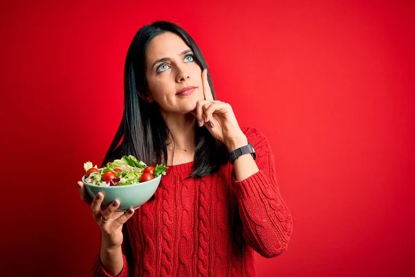 年轻的黑发女人 蓝眼睛 吃着健康的绿色沙拉 而不是孤立的红色背景 严肃的脸在思考问题 非常混乱的想法 — 图库照片