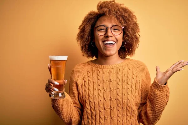 Young beautiful African American afro woman with curly hair drinking glass of beer very happy and excited, winner expression celebrating victory screaming with big smile and raised hands