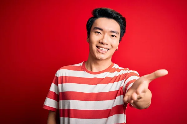 赤い背景に立ってカジュアルな縞模様のTシャツを着て若いハンサムな中国人男性は 挨拶や歓迎として握手を提供フレンドリーな笑みを浮かべて 成功事業 — ストック写真