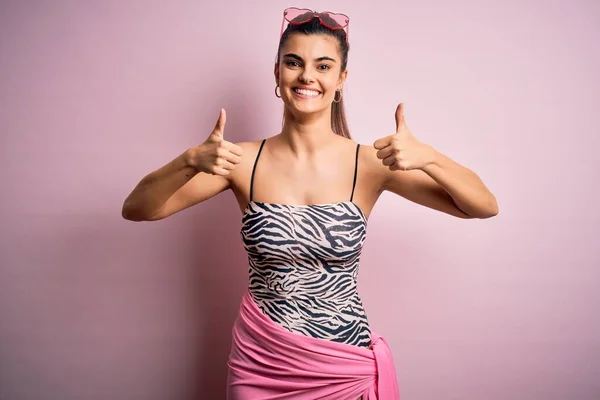 ピンクの背景に水着を着て休暇中の若い美しいブルネットの女性成功サインは手で積極的なジェスチャーをし 親指を上げて笑顔と幸せ 陽気な表情と勝者のジェスチャー — ストック写真