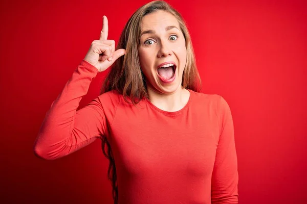 若い美しいブロンドの女性は 成功したアイデアで指を指す孤立した赤い背景の上に立ってカジュアルなTシャツを着ています 興奮し 第一番 — ストック写真