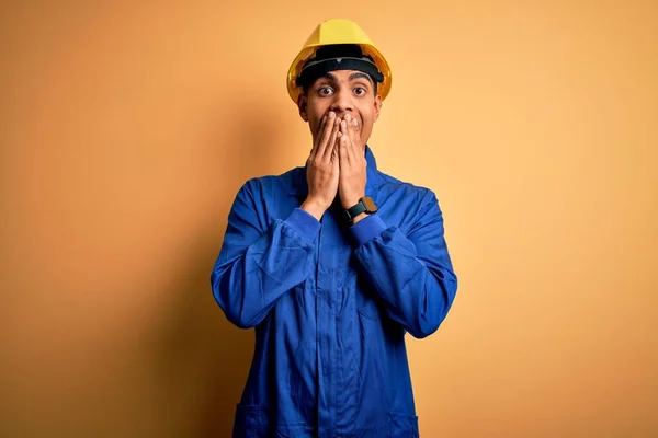 年轻英俊的非洲工人 身穿蓝色制服 头戴安全帽 尴尬地笑着用手捂住嘴 流言蜚语和丑闻的概念 — 图库照片