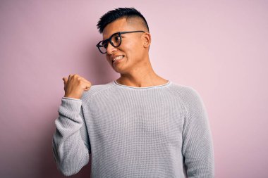 Genç, yakışıklı, Latin bir adam pembe arka planda günlük süveter ve gözlük takıyor. Gülümsüyor, gülümsüyor ve başparmağıyla yan tarafa bakıyor..