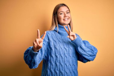 Sarı izole edilmiş arka plan üzerine balıkçı yaka kazak giyen genç, güzel sarışın kadın parmaklarını zafer işareti yaparken gösteren kameraya gülümsüyor. İki numara..