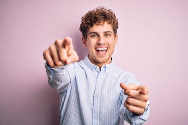 Sarışın, kıvırcık saçlı, beyaz arka planda çizgili tişört giyen, seni ve kamerayı parmaklarıyla gösteren neşeli ve pozitif gülümseyen genç bir adam.