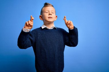 Mavi arka planda mavi süveter giyen, umut ve gözlerin kapalı gülümseyerek el hareketi yapan küçük beyaz çocuk. Şans ve batıl inanç..
