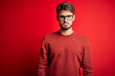 Sakallı, gözlüklü ve süveterli genç bir adam kırmızı arka planda dikiliyor. Depresyonda ve endişeli, sinirli ve korkmuş. Üzücü ifade.
