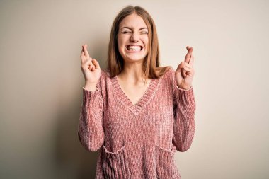 İzole edilmiş beyaz arka planda pembe süveter giyen güzel, kızıl saçlı kadın umut ve gözlerin kapalı bir şekilde gülümseyerek el sallıyor. Şans ve batıl inanç..