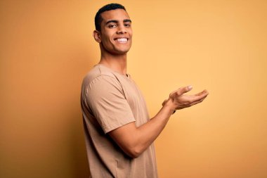 Genç, Afro-Amerikalı, günlük tişörtler giyen, sarı arka planda duran, avuç içleri açık, fotokopi alanı gösteren, mutlu bir gülümseme sunan, genç ve yakışıklı bir adam.