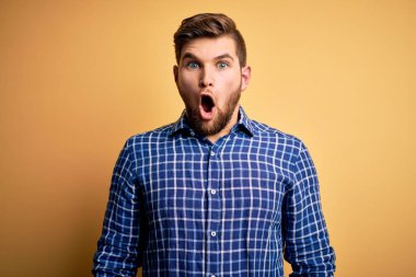 Sarışın, sakallı ve mavi gözlü genç bir işadamı sarı arka planda gömlek giyiyor. Şaşırmış ve şaşırmış bir ifade, korku ve heyecanlı bir yüz ifadesi var..