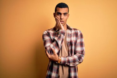 Genç, yakışıklı, Afro-Amerikalı bir adam sarı arka planda dikilmiş, kollarını kavuşturup depresyondan sıkılmış ve yorgun görünüyor..