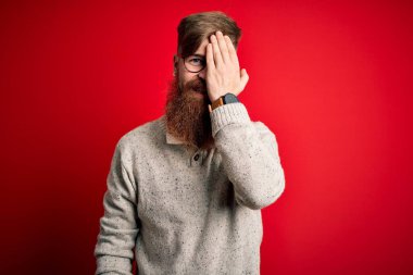 Sakallı, kızıl saçlı, kırmızı arka planda gözlüklü, sıradan bir süveter giyen, bir gözünü kapayan, yüzünde kendine güvenen bir gülümseme ve sürpriz bir duygu olan yakışıklı bir İrlandalı..