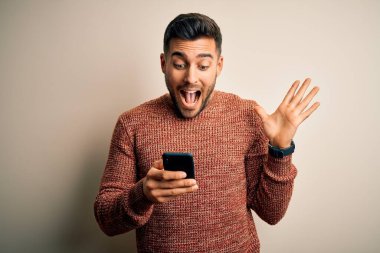 Beyaz arka plan üzerinde akıllı telefon kullanan genç yakışıklı adam çok mutlu ve heyecanlı, kazanan yüz ifadesi zaferi büyük bir gülümsemeyle kutluyor ve ellerini kaldırıyor.