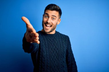 İzole edilmiş mavi arka plan üzerinde duran sıradan süveter giyen genç yakışıklı adam selamlama ve hoş geldin demek için dostça el sıkışma teklif ediyor. Başarılı bir iş..